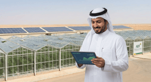 Saudi farmer reviewing energy savings on a tablet with solar panels and greenhouses in the background, illustrating ADF financing opportunities.